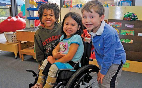Two boys smiling at the camera and standing on either side of a girl in a wheelchair. She is also smiling and looking at the camera.