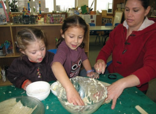 A woman helping two girls mix something in a glass bowl.