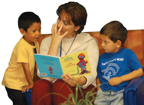 A teacher reading to two boys sitting next to her. The teacher is acting out something from the book by placing her hand in a claw shape near her mouth.