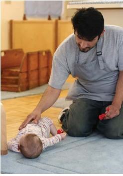 A man sitting next to an infant who is laying on the floor next to him. The man is resting his hand on the baby's stomach and holding a toy.