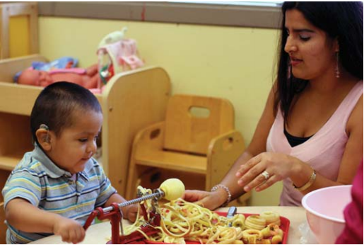 A woman helping a toddler, a deaf boy wearing a striped blue shirt, to make pasta.