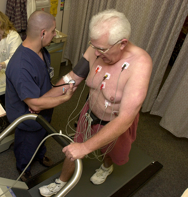 alt="A photo is shown of two men, one walking on a treadmill with various wires connected to his torso region, and the other collecting blood pressure data from the first man."